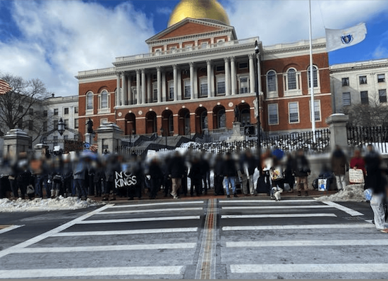 About 20 people lined up in front of the Massachusetts State House. Their faces are blurred.