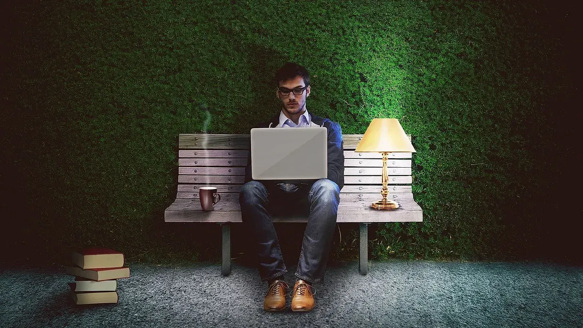 Person in blue jeans and eyeglasses sits alone on a bench, typing on a laptop