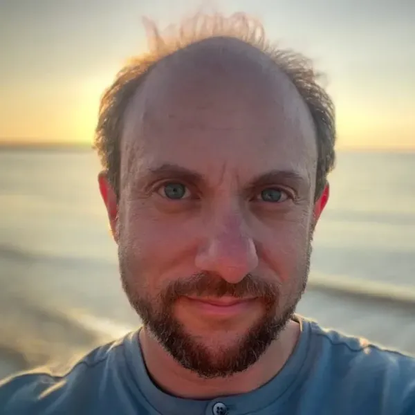 headshot of Tucker Lieberman gazing into camera, hazy ocean in the background, blue T-shirt.