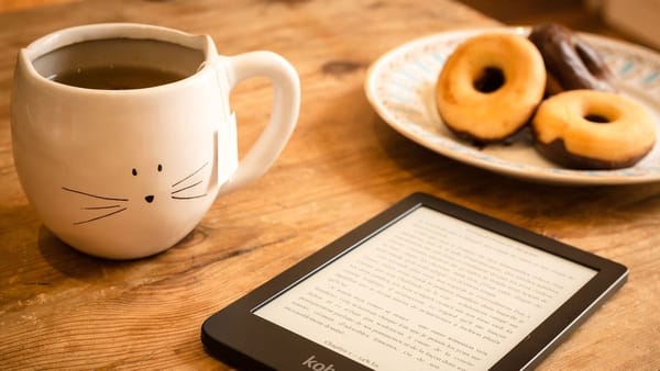 wooden table with a cup of coffee in a mug painted with cat whiskers, a plate of donuts, and an ereader device turned on