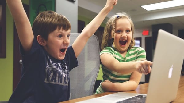 two kids look excited, raising their arms and pointing at a computer screen, as if they've won a video game