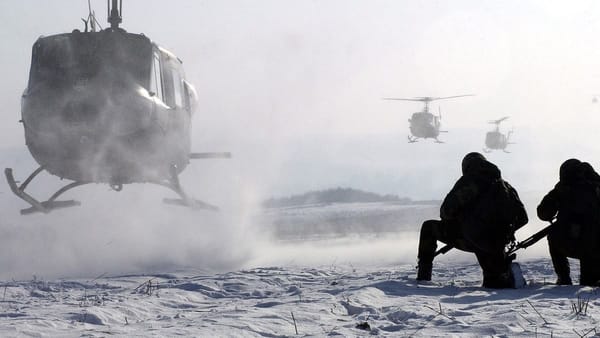 helicopter lands in snow. two people with weapons kneel on the ground. two more helicopters follow close behind.